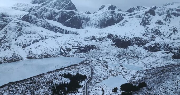 Stortvatnet basin on Austvagoya in Vagan Nordland Norway reveals glacial valley floor with road linking Henningsvaer to inland ridges below Vagekallen massif, drone pullback with upward tilt parallax