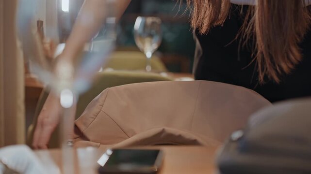 Coat draped over green chair, white guest smoothing fabric reserving seat in cozy bistro wine glasses, phone and bag on wooden table, warm lighting and soft upholstery, hands folding tan jacket
