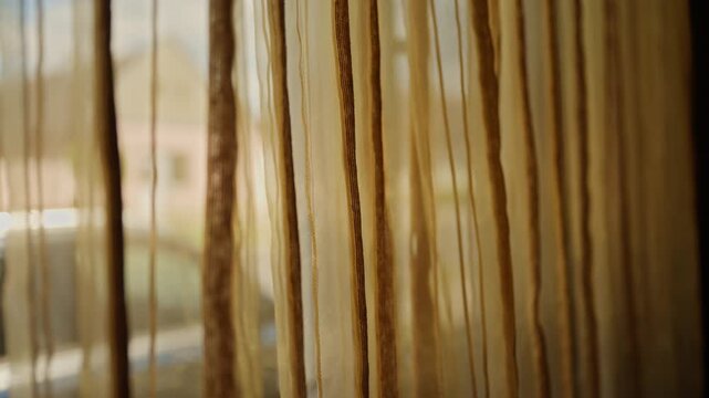 Soft sheer curtains hang at a sunlit window, their vertical textured strands creating a warm, intimate atmosphere and gentle privacy while softly blurring the outdoor view and natural light, 4K.