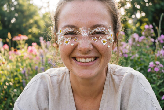 Happy young woman with freckles laughing in summer field wearing creative daisy flower and white lace eye makeup for boho music festival or natural vegan skincare campaign