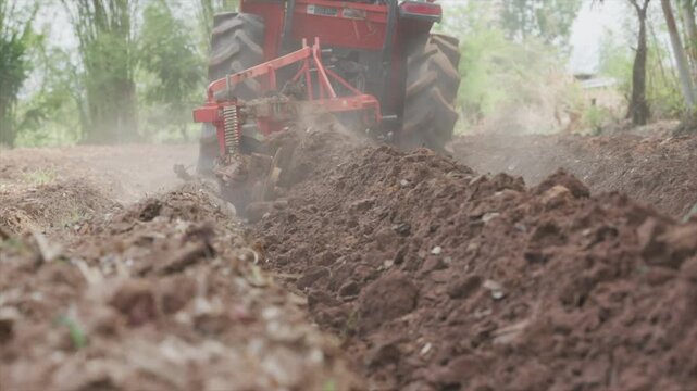 Close-up low-angle shot of a red agricultural tractor pulling a heavy-duty disc plow through rich brown soil on a farm field during a bright sunny day with dust and natural lighting.