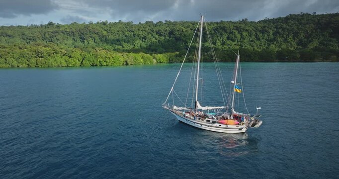 White sailboat anchoring in crystal clear ocean waters next to a lush green tropical island, highlighting a serene scene for travel, adventure, and remote paradise destinations. Aerial view drone shot