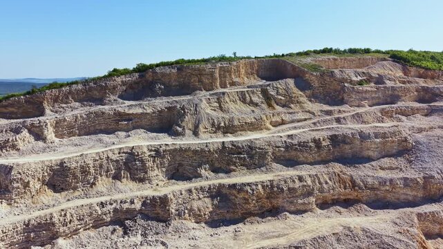A large quarry is shown with distinct layers of rock and dirt. Paths are visible where vehicles operate. The area is surrounded by green bushes under a clear blue sky.