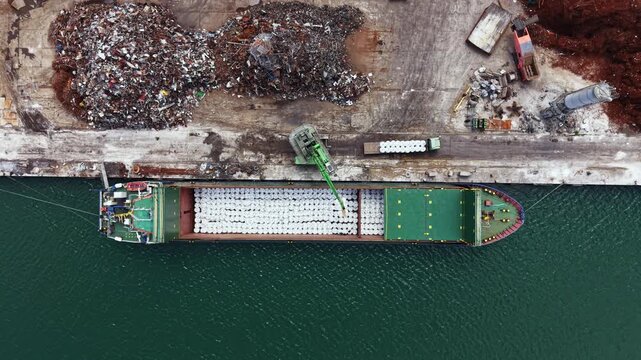 A bulk carrier ship is unloading cargo at a port. A crane is moving materials from the ship to the dock. Various industrial structures are visible in the area.