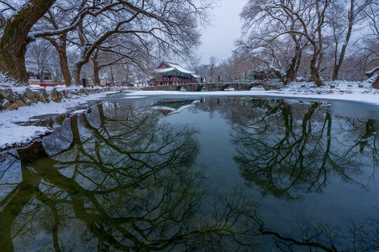 Traditional wooden structure of Gwanghallu Pavilion covered in white winter snow standing beautifully on a frozen pond under an overcast sky in Namwon.