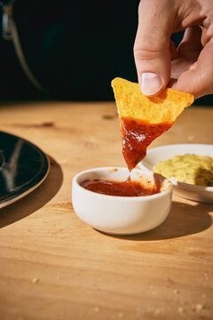 Hand holding tortilla chip coated with red salsa over dipping bowl in closeup. Concept of snack packaging, condiment campaign, restaurant appetizer and food delivery promo.