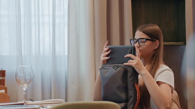 Young white woman unpacking backpack at table, pulling tablet and checking pockets while scanning room, tense expression with eyeglasses, wineglass and plates on table, soft daylight through curtains,