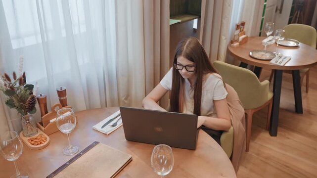 White woman studying on laptop in cafe, taking notes and typing with intent, sunlight filtering through curtains, plant decor and organized table, cup of coffee and textbook nearby, calm bistro