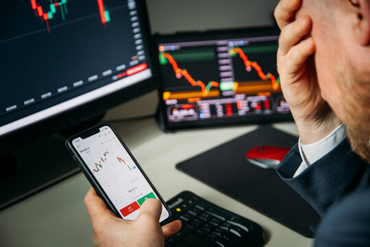 Stressfull trader holding smartphone, using trading app with stock market charts for financial analysis and monitoring investment data.