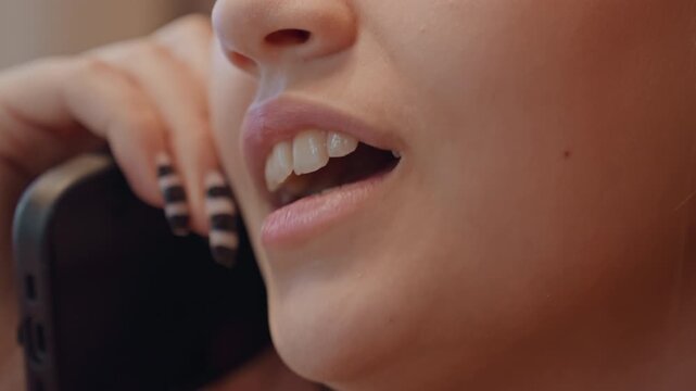 Closeup phone call lips and nails showing smirk and tension young woman arranging client booking and negotiating schedule, whispering confidential detail, striped manicure holding black handset, warm