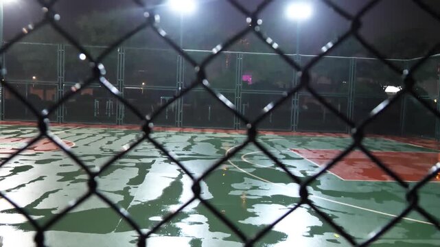 Chain-link fence close-up against a wet, blurry night basketball court with reflections.