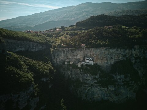 Aerial view of the Sanctuary of Madonna della Corona built into a steep cliffside surrounded by dense green forests and mountain peaks in Italy.