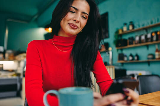 Smiling woman in a red sweater sits in a cozy cafe, scrolling on her phone beside a blue mug