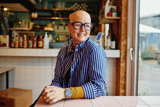Smiling bald woman with glasses relaxed in a cozy cafe wearing striped shirt and smartwatch