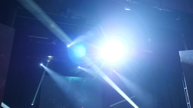 Intense white spotlight beaming from dark ceiling rig creates dramatic blue lens flares and volumetric light rays against black background during professional theatrical concert production.