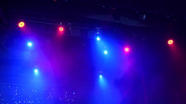 Multiple colored LED spotlights mounted on dark ceiling rig project rotating red and blue light beams through thick theatrical haze during live concert performance.