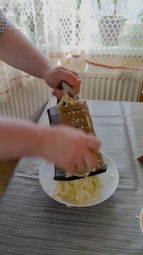 Grating fresh apples on a metal pie grater, Close-up of female hands grating an apple on a classic metal grater into a white plate. Home process of making apple filling or puree in a cozy kitchen