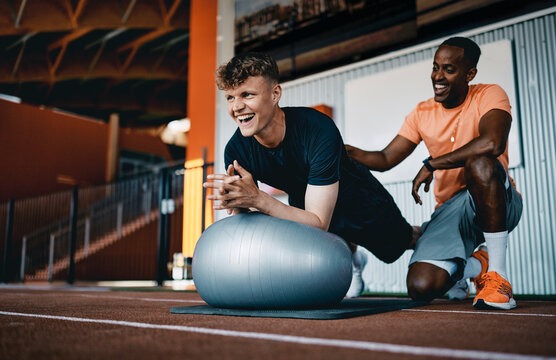 Smiling trainer giving encouragement to a fit young man planking on an exercise ball during a workout session at the gym