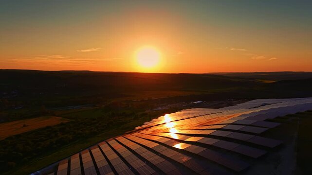 The sun sets in the background, casting orange and yellow colors over a field. Solar panels reflect light, showing the use of renewable energy in an open landscape.