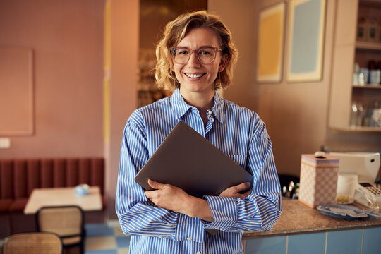 Friendly cafe professional holding a laptop in a cozy cafe, smiling woman with striped shirt