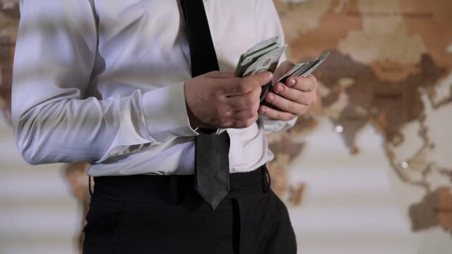 Close-up of a man in a business suit and tie counting a pile of cash dollars in an office against a world map