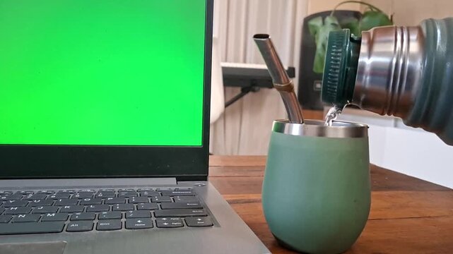 Close up of pouring hot water into a traditional mate cup next to a laptop with a green screen for custom digital content replacement