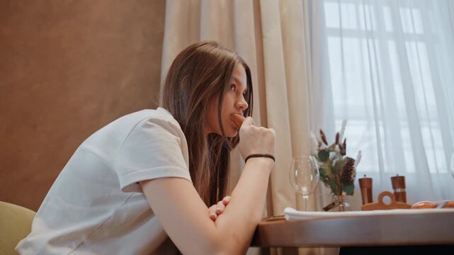 Young black woman sitting at table by window, braided hair holding macaron, contemplative expression, soft sunlight through sheer curtain, warm neutral decor, small pine plant and wine glass on table,