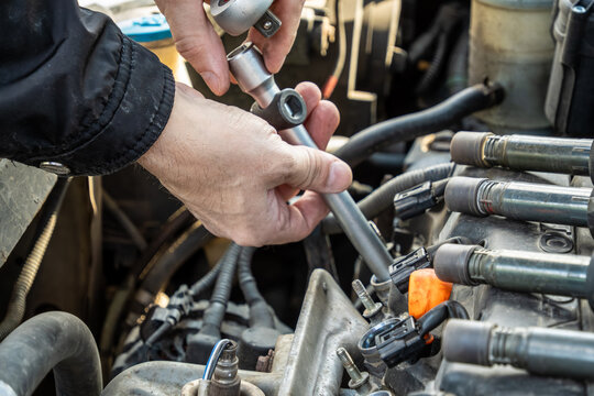 male hands utilizing metal socket ratchet with extension to reach into deep engine cylinder wells for spark plug extraction while disconnected ignition coils lay nearby on top of car motor.