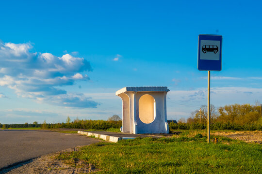 White brutalist-style concrete bus shelter with an oval side opening stands next to an empty asphalt road and a blue bus stop sign, set against a green meadow