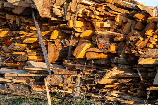 Massive pile comprising irregular sawed timber boards and raw wooden scraps glows under warm evening sunlight showcasing various textures and natural brown hues within a lumberyard environment.