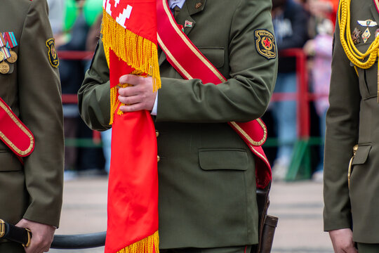 Belarusian officer in olive green parade dress uniform with red sash and shoulder patches holding folded crimson ceremonial banner during national holiday celebration outdoors