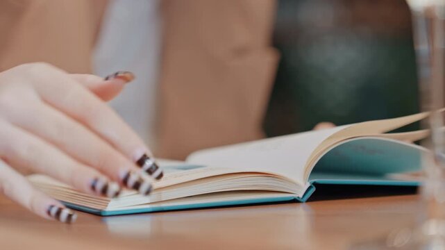 Closeup hands turning book pages, beige blazer, wine glass, gentle manicure, slow journal reading, freelancer sketching ideas, editor proofreading notes, warm bokeh background, peaceful cafe ambiance
