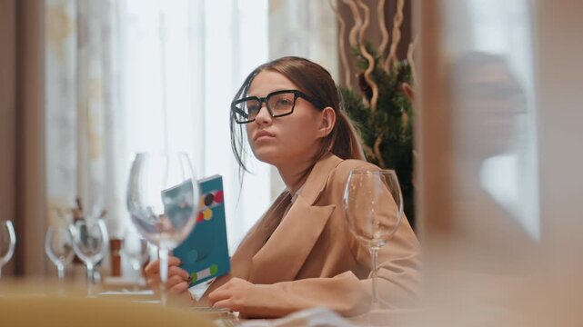 Caucasian woman reading at bistro table, holding colorful lookbook, glasses on, soft window light, empty wine glasses, neutral blazer, focused editorial mood, quiet sophistication