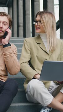 Attractive man and woman sitting on stairs in urban city center working on laptop, talking on mobile phone smiling, stylish freelance people using technology outside, making notes. Outside. Slow