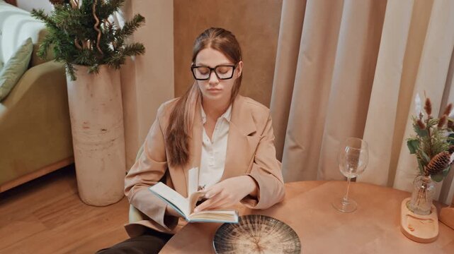 Caucasian woman flipping pages at cafe table, wearing glasses and blazer, thoughtful expression while annotating portfolio, notes and bookmark visible, relaxed posture near curtain and plant, soft
