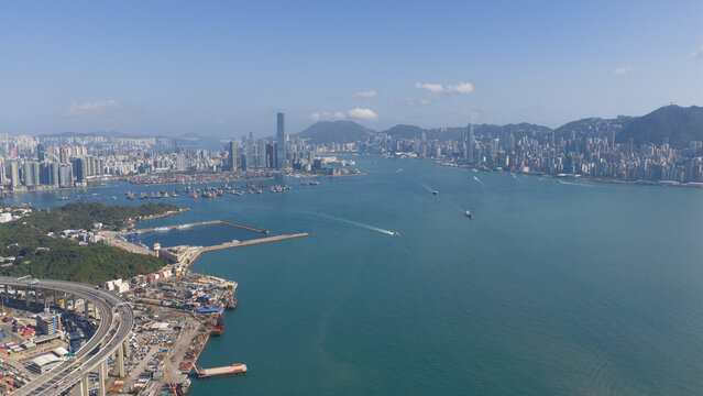 Aerial view of Victoria Harbour with skyscrapers, cargo ships, and a highway bridge under a clear blue sky in Hong Kong, Hong Kong.