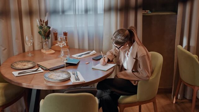Caucasian woman sketching at cafe table with notebook and laptop, coffee cup and pastry plate, bright window light and warm interior, designer tools and color palette on table, focused expression