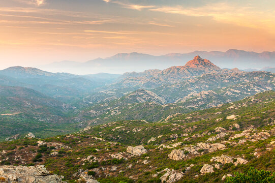 Vast mountain landscape featuring rugged rocky hills and hazy peaks under a vibrant sunset sky, an inspiring travel destination in Agriates desert, Corsica.