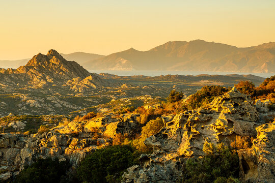 Rugged rocky landscape and distant mountains bathed in golden light, overlooking the sea, a captivating tourism destination in Agriates desert, Corsica.