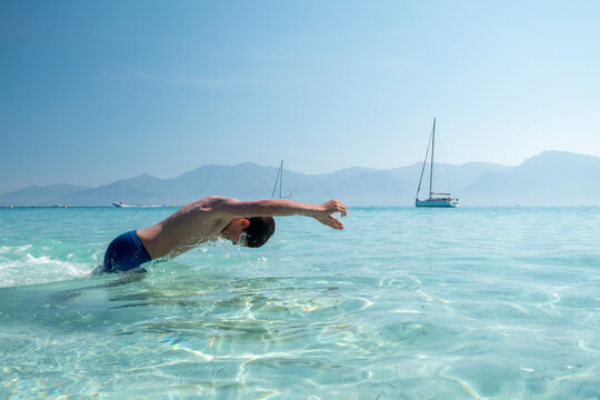 Young boy diving into clear turquoise sea of iconic Saleccia beach with mountains and sailboats, symbolizing summer travel and freedom at Saleccia and Lotu beach, Agriates desert, Corsica.