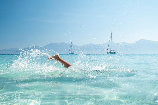 Travel lifestyle: leg splashing in clear turquoise sea with sailboats and distant mountains, symbolizing summer relaxation and freedom at Agriates desert, Saleccia and Lotu beach, Corsica.