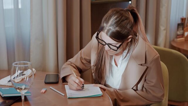 Caucasian consultant writing planner at table organized workspace with glass and phone, natural curtain light, focused productivity, business notes and strategy, client preparation and schedule