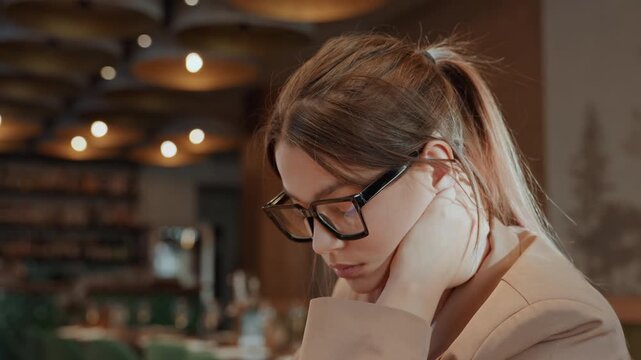 Pensive white woman in cafe, freelancer planning and journaling, ponytail, glasses, elbow on table, bookshelves background, warm lights and bokeh, closed laptop, beverage, relaxed concentration,