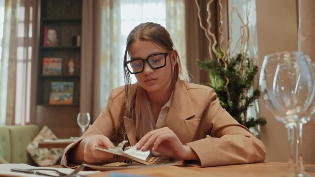 Woman counting cash at cafe table, focused entrepreneur sorting bills and receipts while wearing glasses and trench coat, soft daylight through curtains, elegant interior with wineglass foreground,