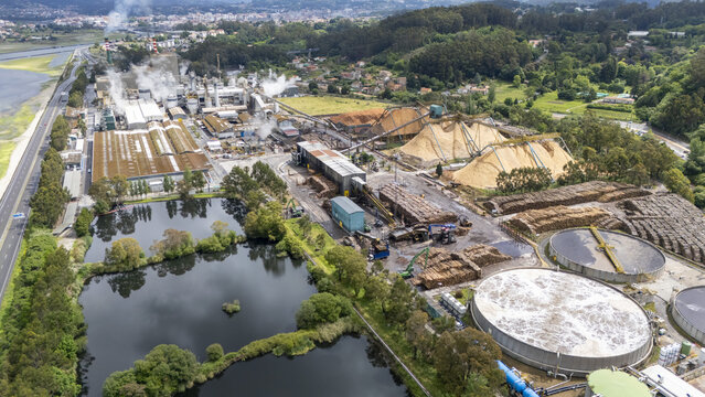 Aerial view of the Biofabrica de Ence pulp mill with large wood chip piles and water treatment tanks surrounded by green hills in Pontevedra, Pontevedra, Spain.
