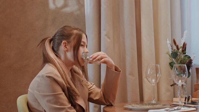 Young woman staring out near table, leftover plates and decorative vase with seasonal greenery, soft warm interior light, small festive tree in corner, introspective and pensive atmosphere, intimate