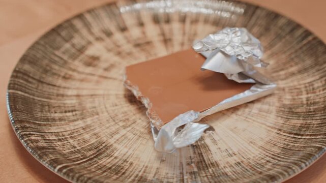 Empty ceramic plate before chocolate appears, hand reaches in to place partially wrapped milk chocolate, foil crumples, textured plate pattern, warm surface, cozy snack preparation, closeup sequence,