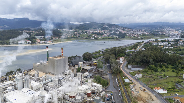Aerial view of the Biofabrica de Ence industrial complex with smoking chimneys near the river estuary and green hills in Pontevedra, Pontevedra, Spain.