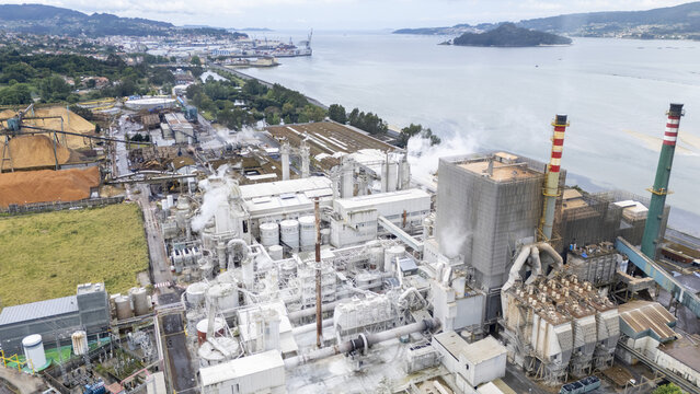 Aerial view of the Biofabrica de Ence pulp mill with industrial structures, smoke rising from chimneys, and wood chip piles along the coast in Pontevedra, Pontevedra, Spain.