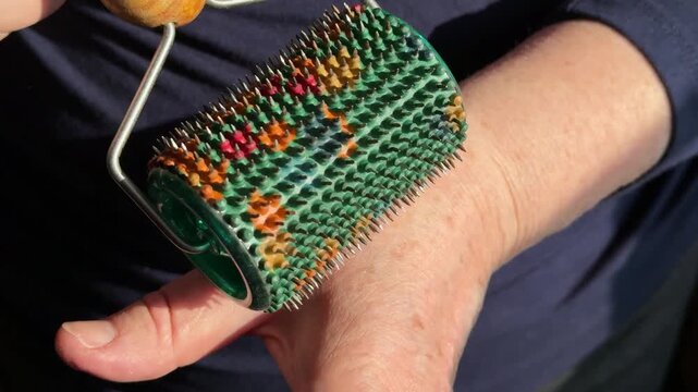 Close-up woman hands rolling green needle roller, wooden handle guiding colorful spikes across skin, slow rhythmic motion